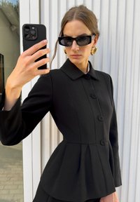 Žena v černé zapínací peplum bundě, s velkými zlatými kruhovými náušnicemi, černými obdélníkovými slunečními brýlemi, jak si dělá selfie s černým chytrým telefonem.