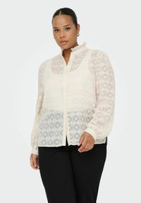 Blusa leggera e trasparente color crema con motivi geometrici, colletto alto e chiusura con bottoni. Maniche lunghe con polsini arricciati.