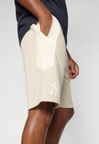 Beige shorts med strukturert stoff, med en kontrasterende hvit lomme og trykt logo på kanten. Designet inkluderer sidelommer.