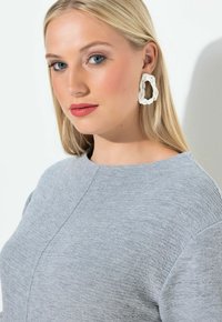 Šedý texturovaný top s kulatým výstřihem, doplněný velkými bílými vlnitými náušnicemi. Hladké vlasy, jemný make-up a neutrální pozadí.