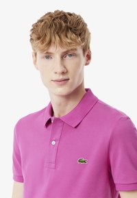 Kortärmad polo i ljusrosa, tillverkad av strukturerad bomull, med en tvåknappsknäppning och en grön Lacoste-logotyp på bröstet.