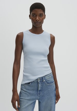 Femme aux cheveux courts portant un haut côtelé sans manches bleu clair et un jean bleu taille haute sur un fond uni.