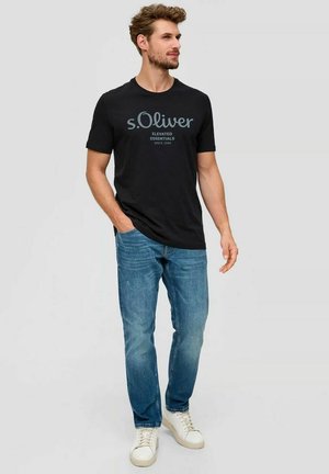 Man wearing black s.Oliver logo t-shirt, blue jeans, and white sneakers, standing with one hand in pocket, looking right.