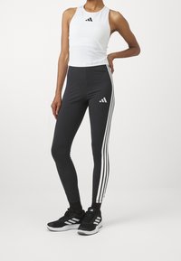 Personne portant un haut sans manches blanc, des leggings noirs avec des bandes blanches sur les côtés, des chaussures de sport noires, posant avec une main sur la hanche.