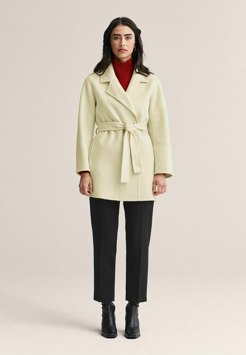 Krämfärgad kappa med avtagbart bälte i midjan, kragen, och långa ärmar, tillsammans med svarta byxor och ankelstövlar. Röd turtleneck under.