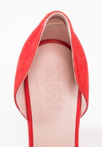 Chaussure plate ouverte en rouge daim avec une semelle intérieure beige clair embossée du nom de la marque "ZIGN", visible de la vue de dessus.