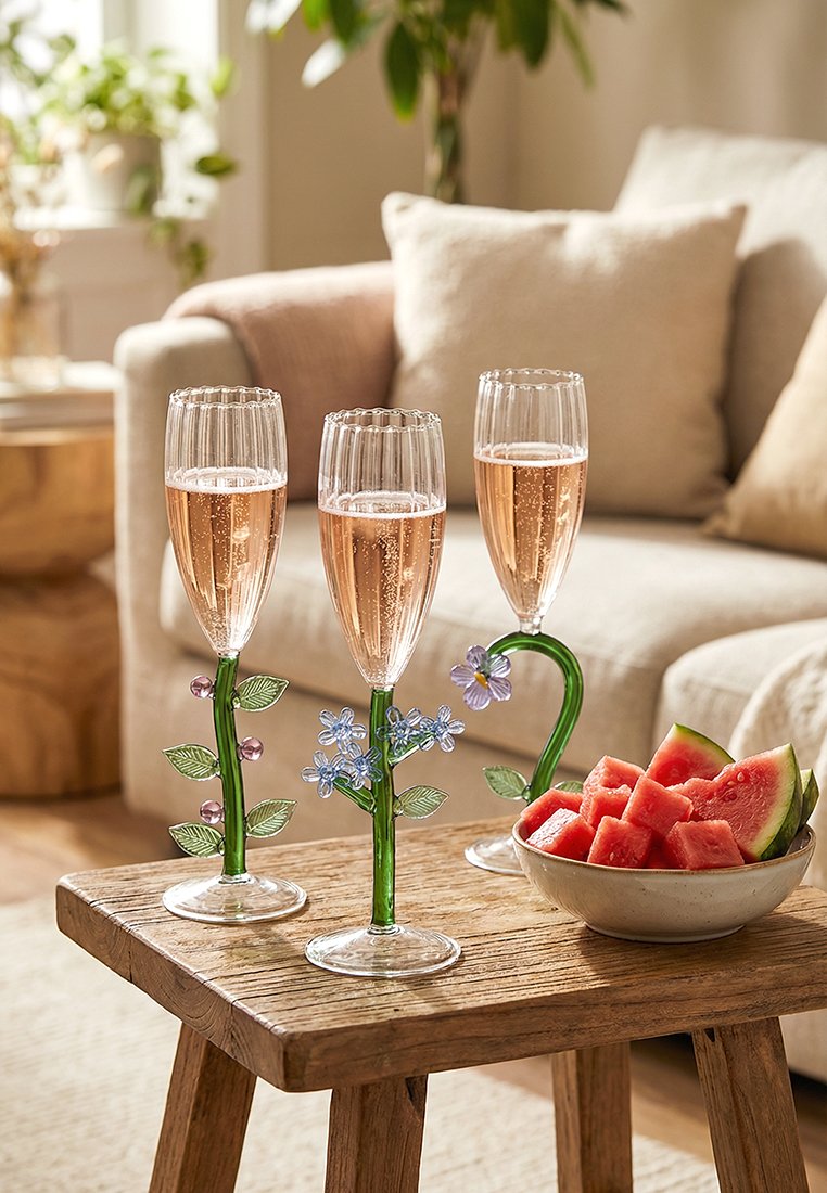 Trzy wysokie kieliszki do szampana z kwiatowymi trzonkami, wypełnione musującym różowym winem, stojące na drewnianym stole obok miski z plasterkami arbuza.