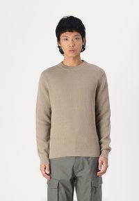 Long-sleeve beige sweater with ribbed texture, round neck, fitted cuffs, and a straight hem, paired with olive-green cargo pants.