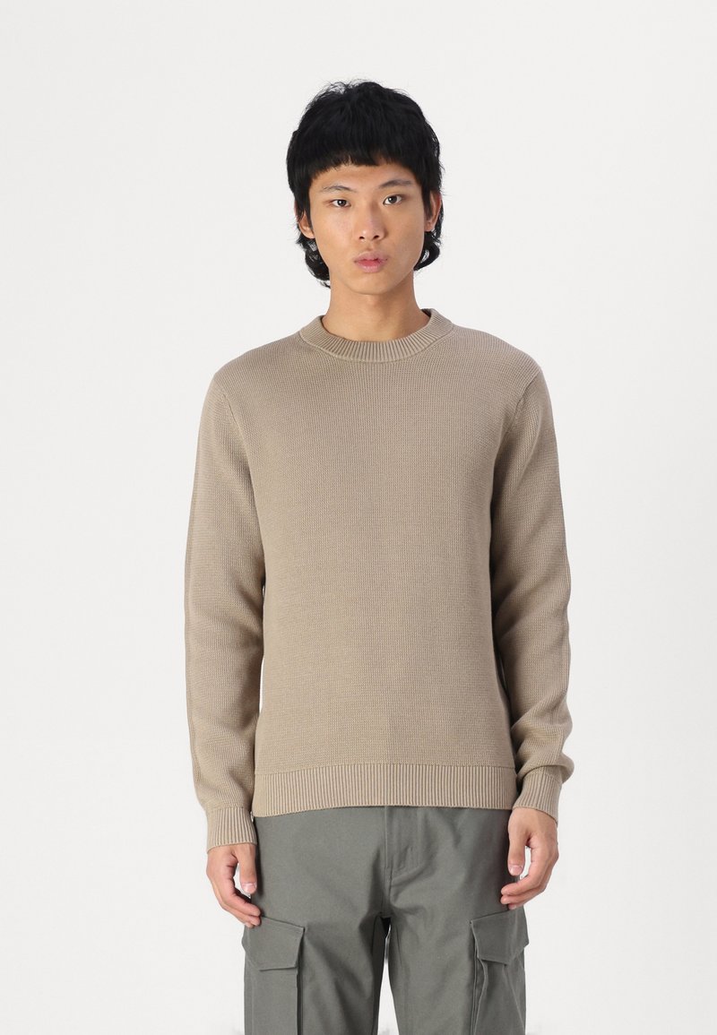 Long-sleeve beige sweater with ribbed texture, round neck, fitted cuffs, and a straight hem, paired with olive-green cargo pants.