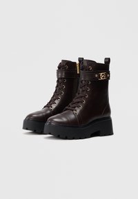 Stivaletti in pelle marrone con hardware dorato, design con lacci, suola larga e chunky, e cerniera laterale per una facile vestibilità.