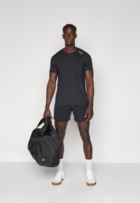 Zwarte sport T-shirt met ronde hals, gecombineerd met zwarte shorts. Het model houdt een zwarte sporttas vast. Witte sportschoenen en sokken zijn gedragen.