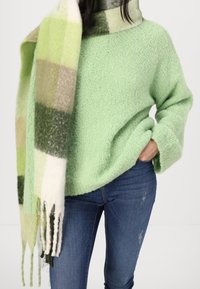 Pull vert clair texturé à coupe ample avec manches retroussées, associé à une écharpe à carreaux dans des tons verts, beiges et crème. Jean foncé visible.