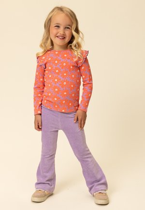 Oranje shirt met lange mouwen, een bloemmotief en gerimpelde mouwen, gecombineerd met paarse corduroy uitlopende broeken en beige sneakers.