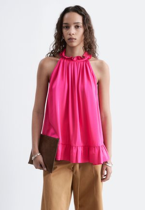 Jeune femme aux cheveux ondulés portant un haut sans manches rose vif à volants et un pantalon beige, tenant une pochette marron foncé.