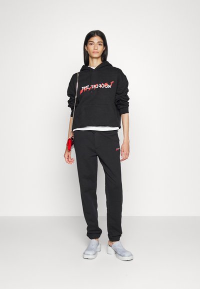 Schwarze, cropped Kapuzenjacke mit rotem grafischen Text, kombiniert mit schwarzen Sweatpants und grauen Slippern. Elegante, lässige Sportbekleidung.