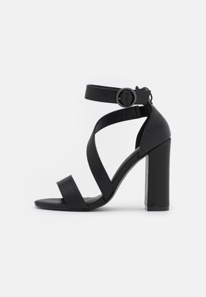 Sandale noire à talons hauts avec un large talon bloc, une large bride avant, des brides croisées au milieu, et une bride de cheville avec boucle ronde sur fond blanc.