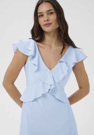 Žena s dlhými tmavými vlasmi v svetlomodrých šatách s volánikovými rukávmi a hlbokým výstrihom do V, stojaca pred jednoduchým pozadím.