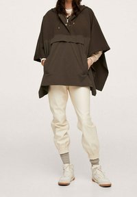 Poncho à capuche marron foncé avec deux poches avant, associé à un pantalon de jogging couleur crème et des baskets blanches. Tissu lisse, coupe décontractée.