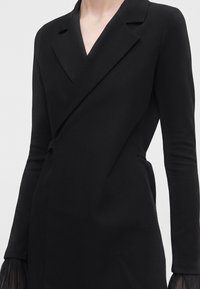 Femme portant une robe noire cintrée à manches longues avec un col en V profond et des poignets plissés semi-transparents sur un fond clair uni.