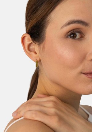 Frau mit braunen Haaren, die kleine goldene Kettenglied-Ohrringe trägt, legt die Hand auf die Schulter und schaut leicht seitlich von der Kamera weg vor weißem Hintergrund.