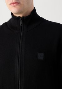 Svart zip-up tröja gjord av mjukt material, med hög krage, ribbade detaljer och en liten präglad logotyp i övre vänstra hörnet.