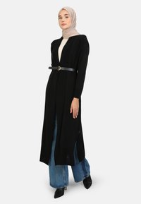Zwarte lange cardigan met een riem in de taille, voorzien van zijsplitten, gedragen over een lichte top en gecombineerd met een brede spijkerbroek en hoge schoenen.