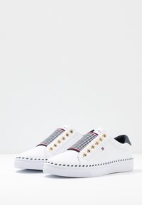 Baskets en toile blanche avec bande élastique rayée, œillets dorés et accents noirs. Semelle en caoutchouc avec bordure bleu marine. Sans lacets.