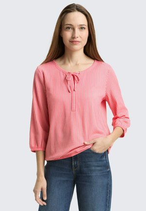 Frau mit langen braunen Haaren, die eine rosa-weiß gestreifte Bluse mit 3/4-Ärmeln trägt, die am Hals gebunden ist, und blaue Jeans, steht mit einer Hand in der Tasche.