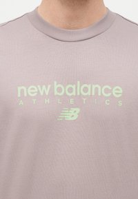 Κοντινό πλάνο ενός μωβ μπλουζακιού με στρογγυλή λαιμόκοψη, με ανοιχτό πράσινο κείμενο "new balance ATHLETICS" και το λογότυπο New Balance στο στήθος.