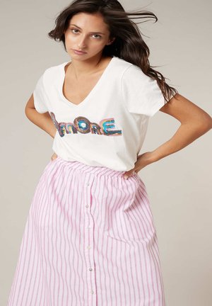 Femme aux longs cheveux bruns portant un T-shirt blanc à col en V avec le texte coloré "Amore" et une jupe taille haute rayée rose et blanche avec des boutons.