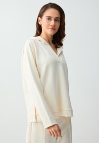 Jeune femme aux cheveux bruns portant un sweat à capuche crème ample et un pantalon assorti, debout devant un fond gris clair uni.