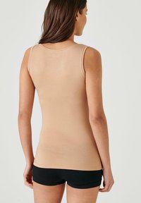 Nude-colored, form-fitting tank top, with thick straps and a smooth texture. Worn with black shorts, showing a simple, classic design.