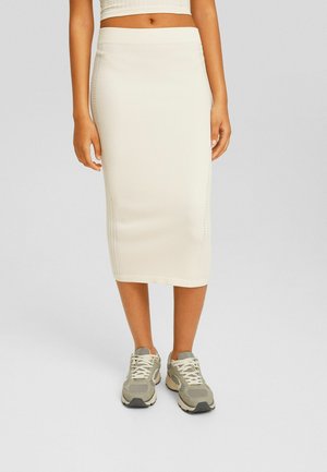 Vrouw met een witte geribbelde midi-rok en beige-grijze veter sneakers, staand tegen een effen achtergrond.