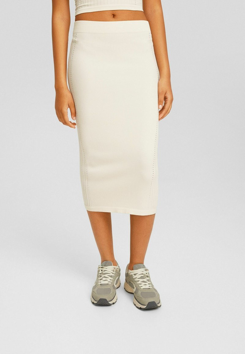 Femme portant une jupe midi blanche côtelée et des baskets beige-gris lacées, debout devant un fond uni.