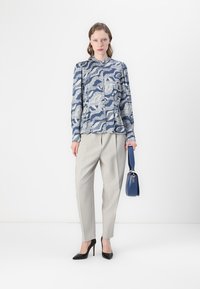 Blue patterned blouse with long sleeves paired with light grey pleated trousers. Model holds a blue handbag and wears black stiletto heels.