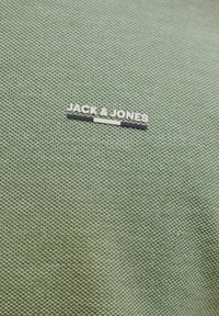 Tejido verde texturizado con un patrón tejido; incluye una etiqueta de logo plateado que dice "JACK & JONES" con acentos en negro y blanco.
