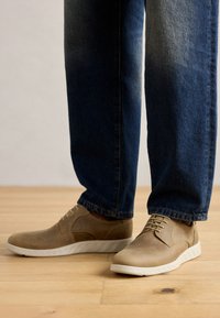 Brune-suede sko med rund tå og hvid gummisål, parret med mørkeblå jeans med lige ben. Baggrund i naturligt trægulv.