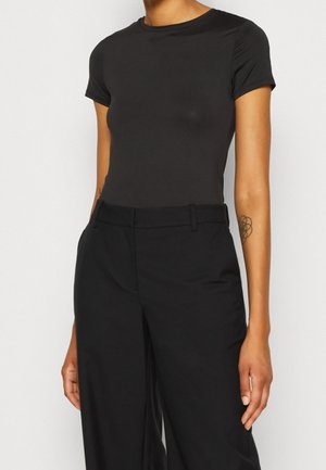 Personne portant un t-shirt noir ajusté à manches courtes rentré dans un pantalon large noir taille haute, sur un fond uni.