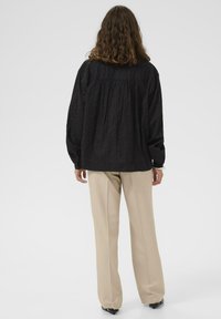 Frau mit schulterlangen, lockigen Haaren, die eine lockere schwarze Bluse und beigen, weiten Hosen trägt, steht mit dem Rücken zu einer weißen Wand.