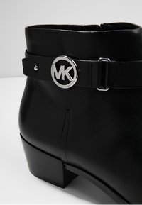 Bottine noire en cuir avec un petit talon carré, dotée d'une bride décorative et d'une boucle argentée avec le logo MK de Michael Kors.