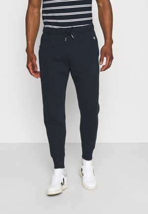 Mann mit schwarzen Jogginghosen mit Kordelzug am Bund, weißen Turnschuhen und einem schwarz-weiß gestreiften Shirt, stehend auf einem grauen Boden.