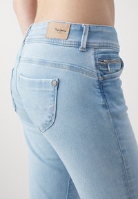 Svjetloplave traperice s patentnim zatvaračem, zakrivljenim stražnjim džepovima i kožnom etiketom s natpisom "Pepe Jeans" na pojasu.