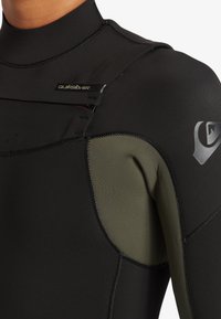 Čierny neoprénový oblek s tesným strihom, s zelenými akcentmi po stranách, textúrovaným povrchom a hladkými švami. Logo na hornej ľavej časti hrude.