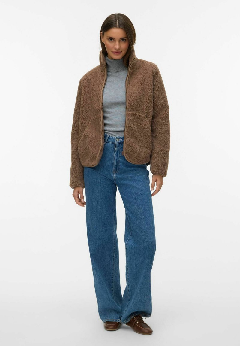 Femme portant une veste en polaire marron, un pull à col roulé gris, un jean large bleu et des chaussures marron, debout devant un fond blanc.