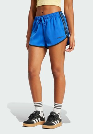Pantaloni sportivi blu con dettagli neri, dotati di un cinturino con cordino, fessure laterali e logo. Abbinati a sneakers bianche e nere.