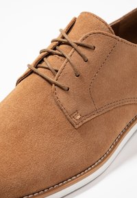 Chaussure en daim marron avec une texture lisse, dotée d'un design à lacets traditionnel et d'une semelle en caoutchouc blanche avec un accent beige.