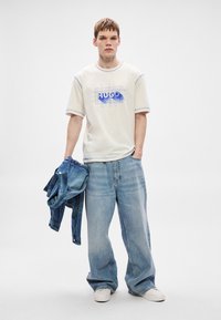 Camiseta branca com estampa azul "HUGO", combinada com jeans largos de ganga azul-claro. Um casaco de ganga está pendurado sobre um dos braços.