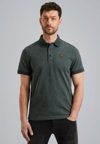 Mørkegrøn polo-shirt med et struktureret m�ønster, to-knaps knaplukning og et broderet logo på brystet. Korte ærmer og kravedesign.