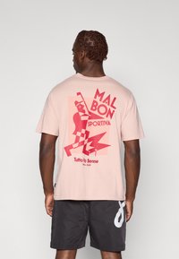 Lyserød bomulds t-shirt med en rød grafisk design med teksten "MAL BON SPORTIVA" og geometriske mønstre på ryggen. Korte ærmer.