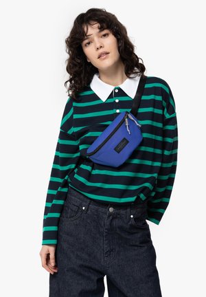 Blå textilaxelväska med dragkedja, bärs över en svart och grön randig tröja, i kombination med mörk denimbyxor.